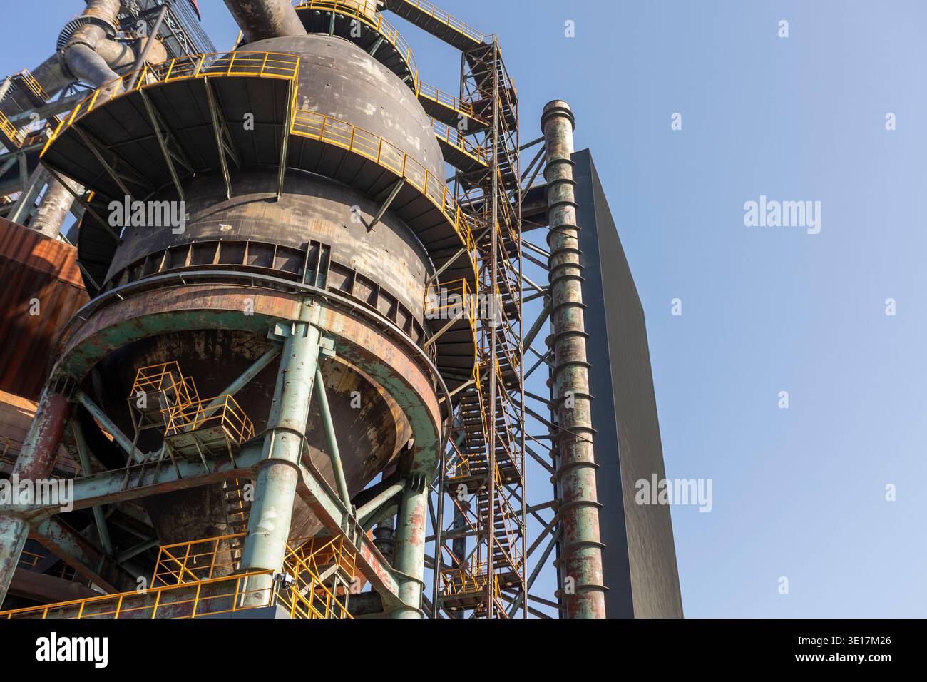 Una torreggiante struttura industriale in metallo con superfici arrugginite e ringhiere gialle è posta su un cielo blu limpido, catturando l'essenza della metallurgia Foto Stock