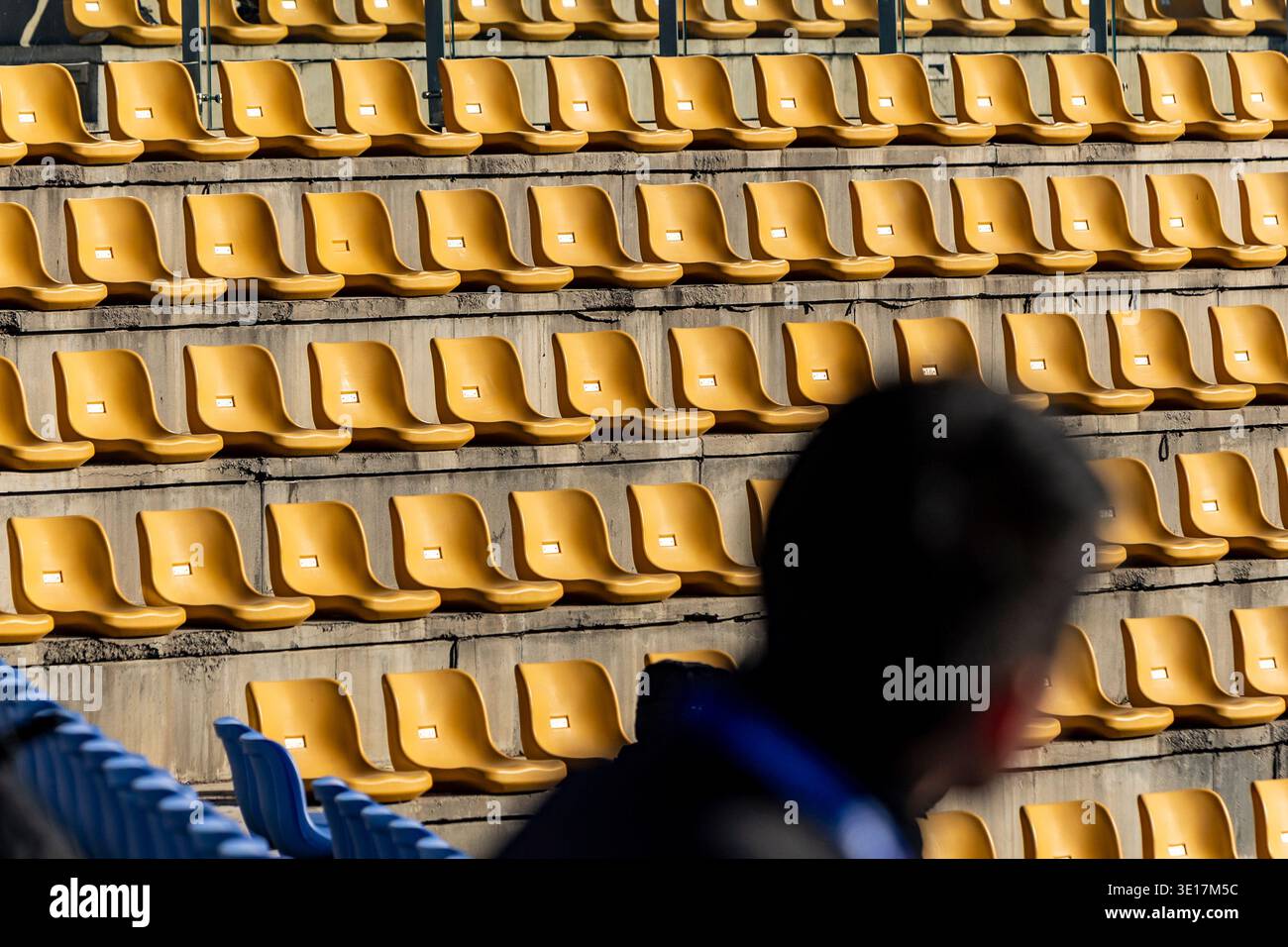 File di sedie da stadio gialle vuote crogiolano alla luce del sole, gettando ombre e creando un modello di ripetizione. Una figura sfocata si trova in primo piano, Foto Stock