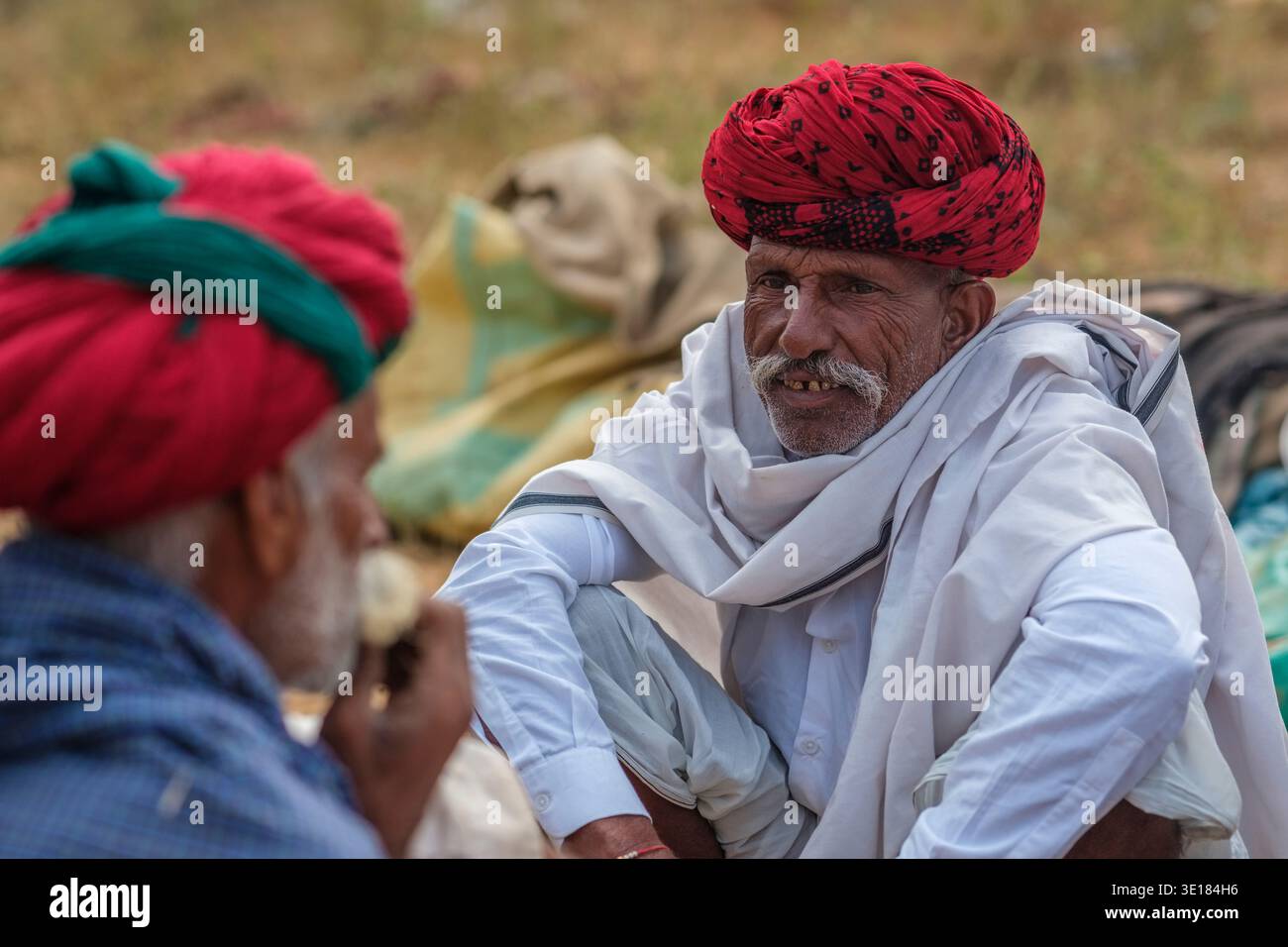 Alla Fiera dei cammelli di Pushkar, un uomo con un turbante dai motivi rossi e abiti bianchi si impegna in una conversazione mentre è seduto sul terreno sabbioso. Foto Stock