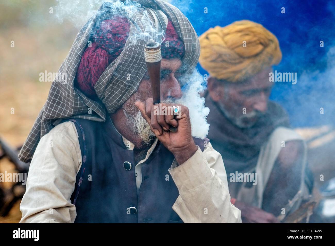 Un uomo in un turbante controllato espira fumo mentre usa una pipa tradizionale alla Fiera dei cammelli di Pushkar con un altro uomo seduto a distanza nebulosa. Foto Stock