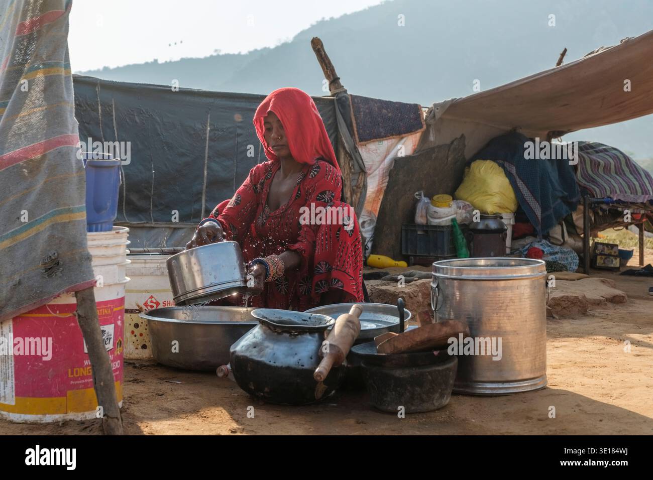 Una donna con un indumento rosso lava piatti metallici in un campo improvvisato durante la Fiera del cammello di Pushkar con una grande collina sagomata in lontananza. Foto Stock