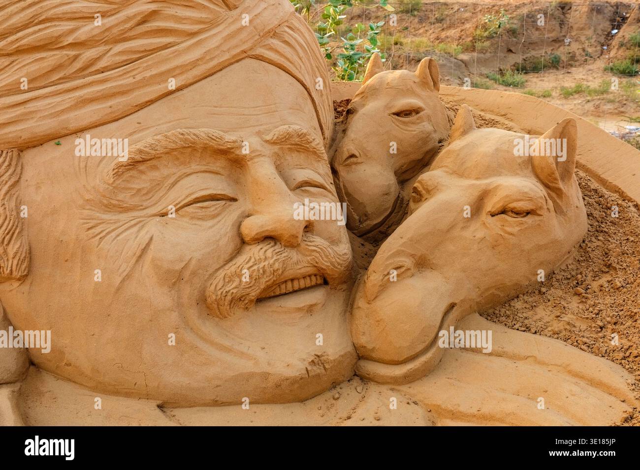 Primo piano di una scultura di sabbia alla Fiera dei cammelli di Pushkar, che mostra un uomo sorridente in un turbante accanto alle teste dettagliate di due cammelli. Foto Stock