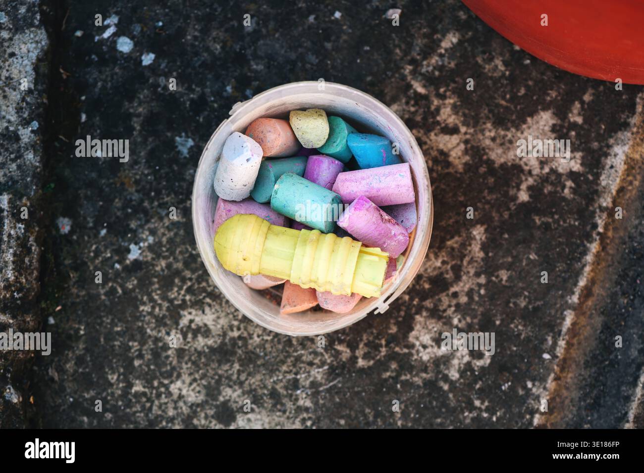 Vista dall'alto dei colorati pezzi di gesso del marciapiede in un contenitore di plastica su cemento grezzo all'aperto, vivaci tonalità pastello che contrastano con il surf intempestivo Foto Stock