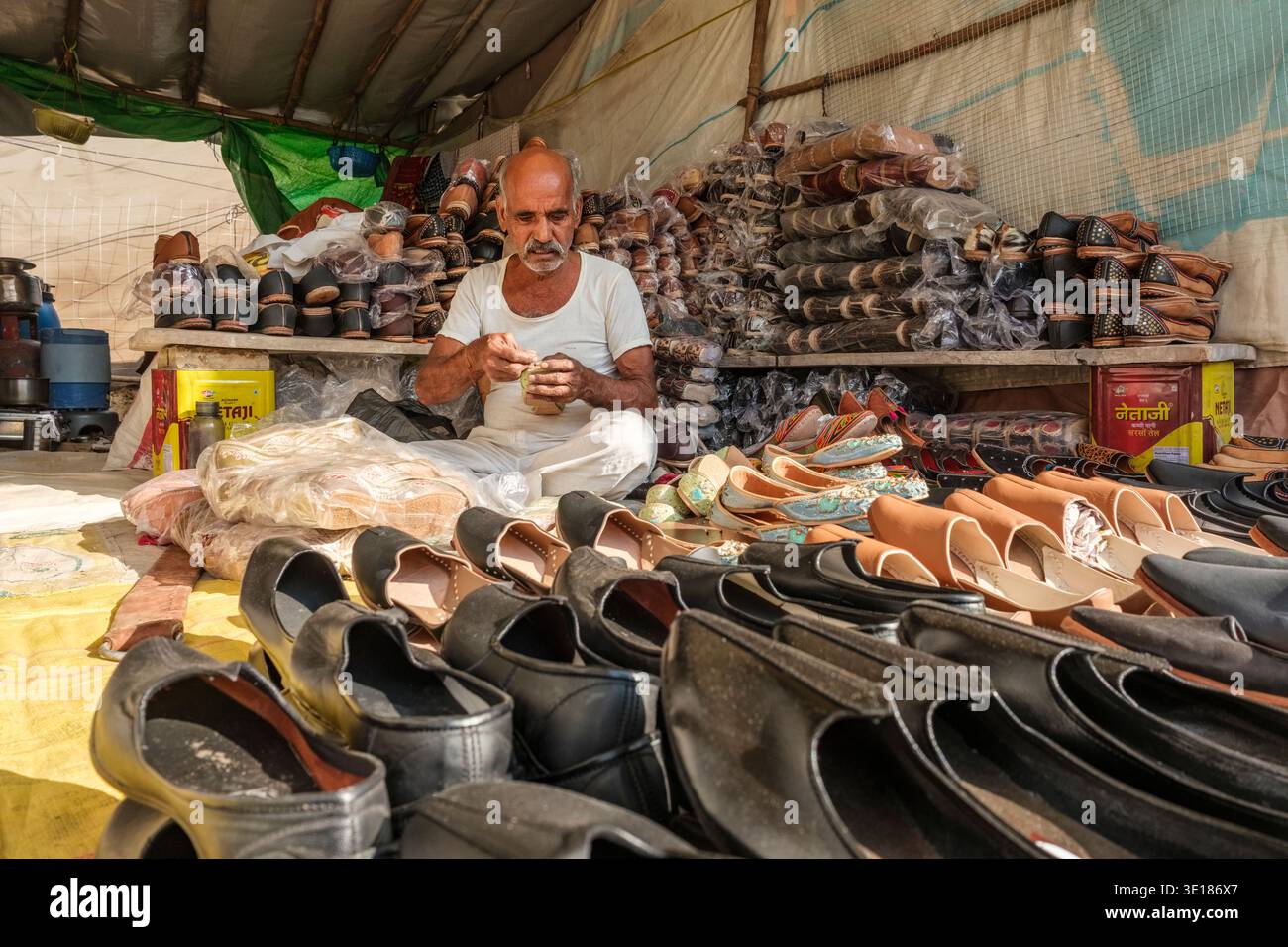 Un anziano artigiano siede tra file di calzature tradizionali in pelle a Pushkar Mela, cucendo con cura una scarpa all'interno del suo laboratorio temporaneo. Foto Stock