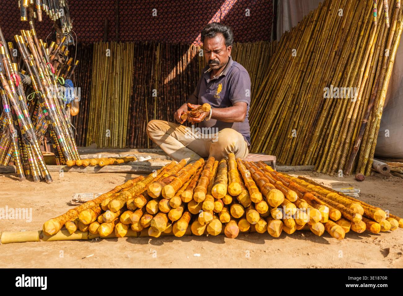 Un venditore al Pushkar Mela lucida meticolosamente il lungo personale di legno seduto a terra circondato da pile di bambù grezzo e finito. Foto Stock