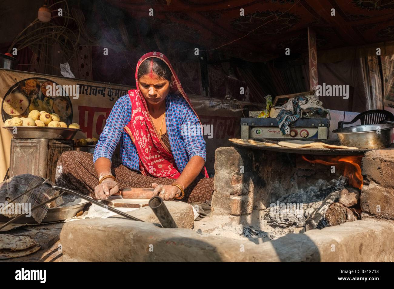 Una donna prepara la focaccia tradizionale sopra un caminetto aperto in una bancarella di cibo di Pushkar Mela, con pile di spuntini cotti visibili in lontananza. Foto Stock
