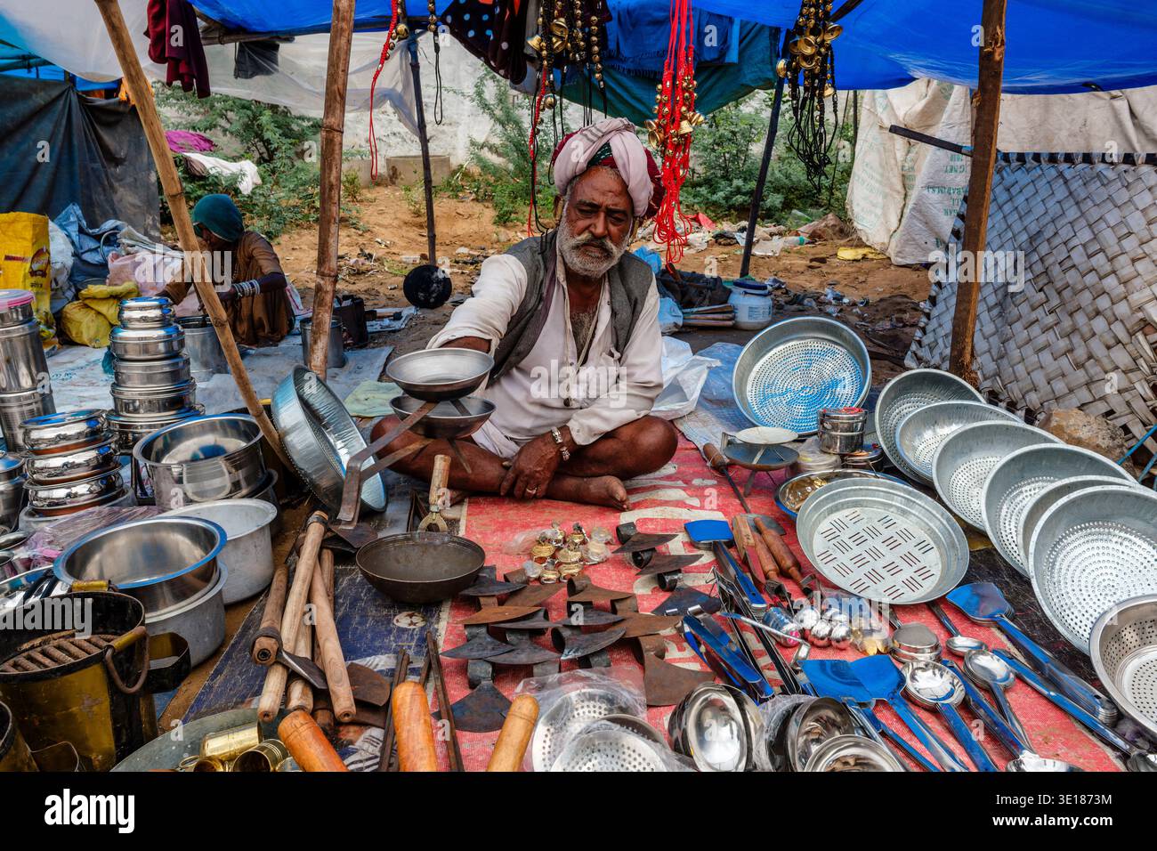 A Pushkar Mela, un mercante riposa dietro una serie di utensili da cucina in metallo e utensili in ferro disposti su un panno rosso sotto un semplice telone in tessuto blu. Foto Stock