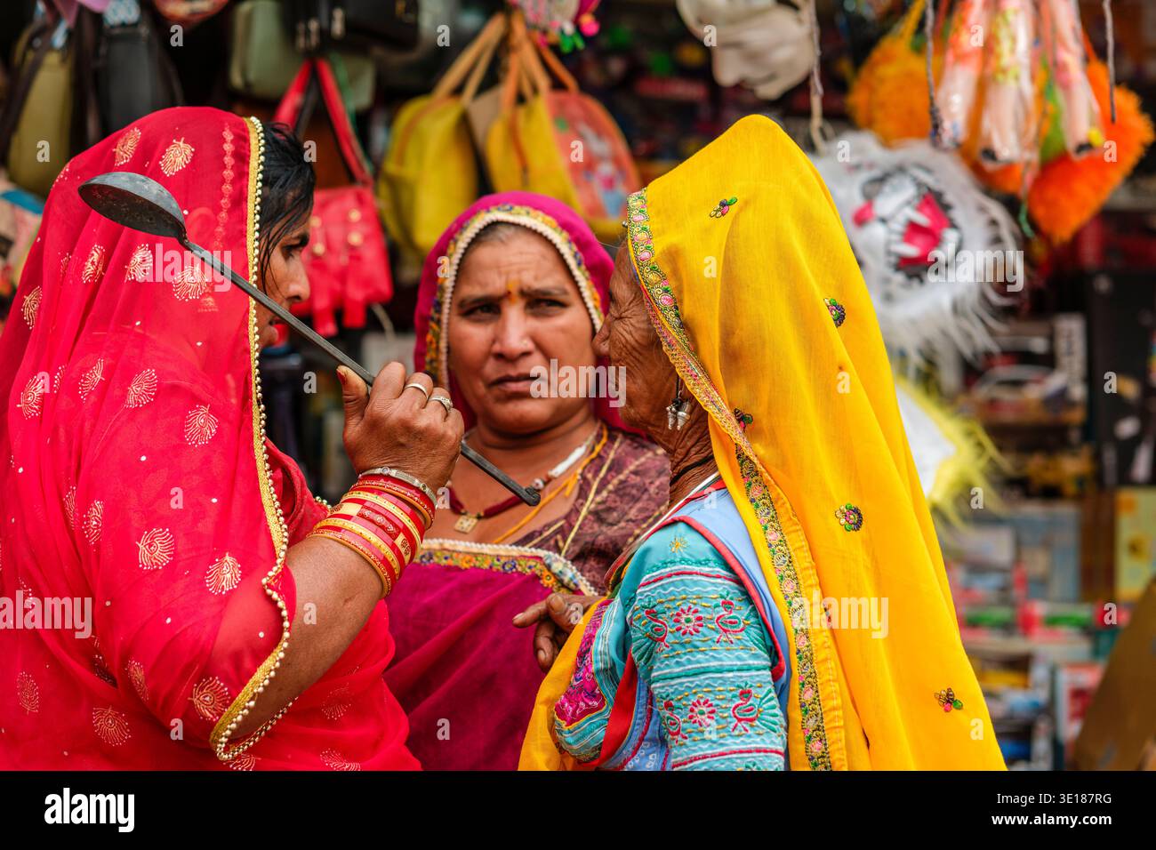 Un gruppo di donne in saris ricamati a Pushkar Mela si trova vicino a un negozio di souvenir, con una che tiene un mestolo di metallo mentre altre parlano intensamente. Foto Stock