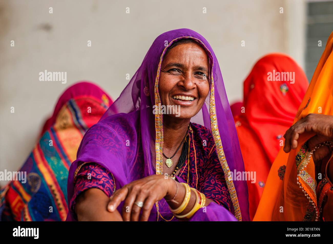 Una donna sorridente in un sari viola e bracciali gialli si siede con altre persone vestite in modo brillante a Pushkar Mela. Foto Stock