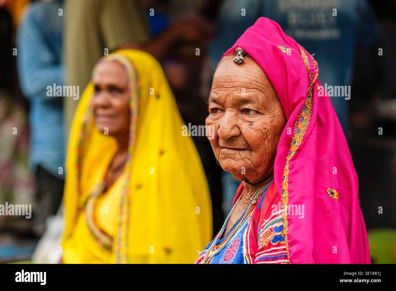 Una donna anziana a Pushkar Mela che indossa un velo rosa guarda di lato mentre una donna di colore giallo si siede dietro di lei. Foto Stock