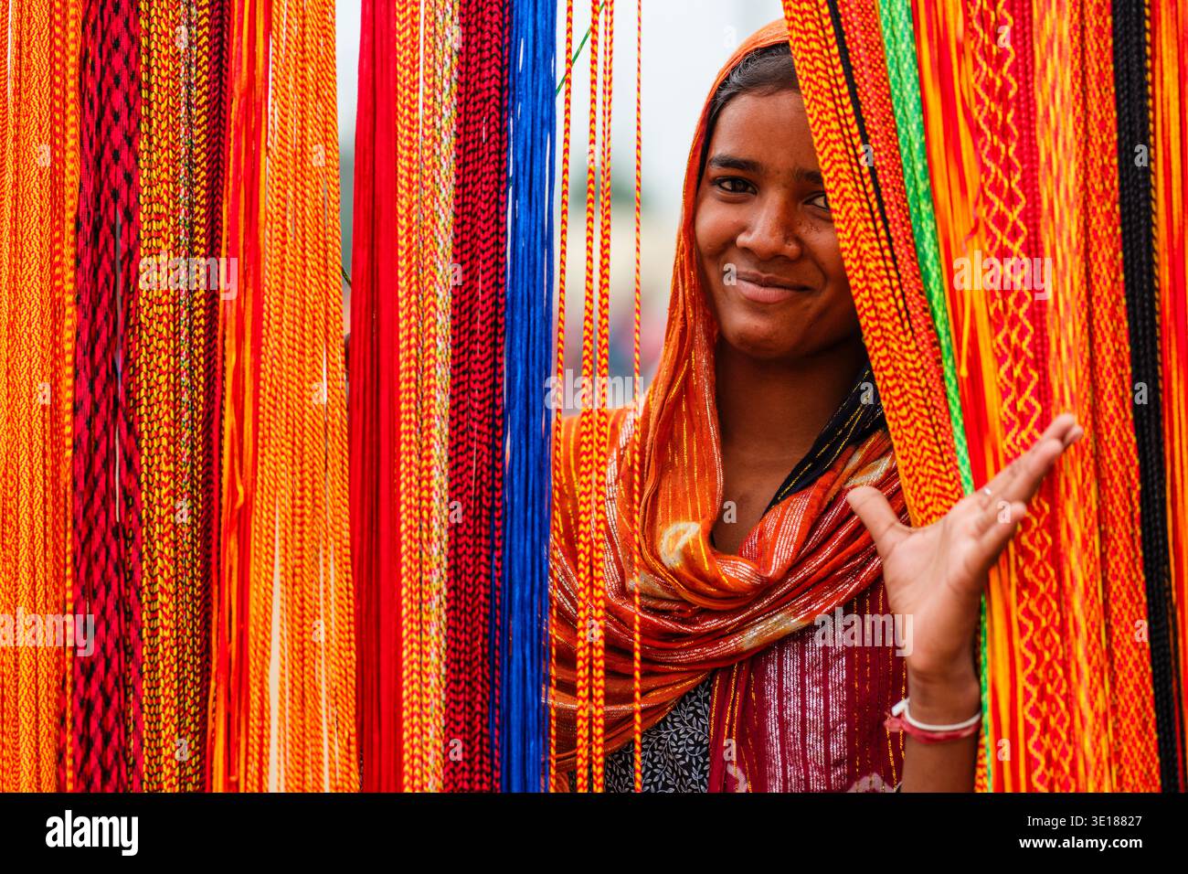 Una giovane donna sorridente di Pushkar Mela si posa dietro una fitta cortina di corde intrecciate rosse, gialle e blu. Foto Stock