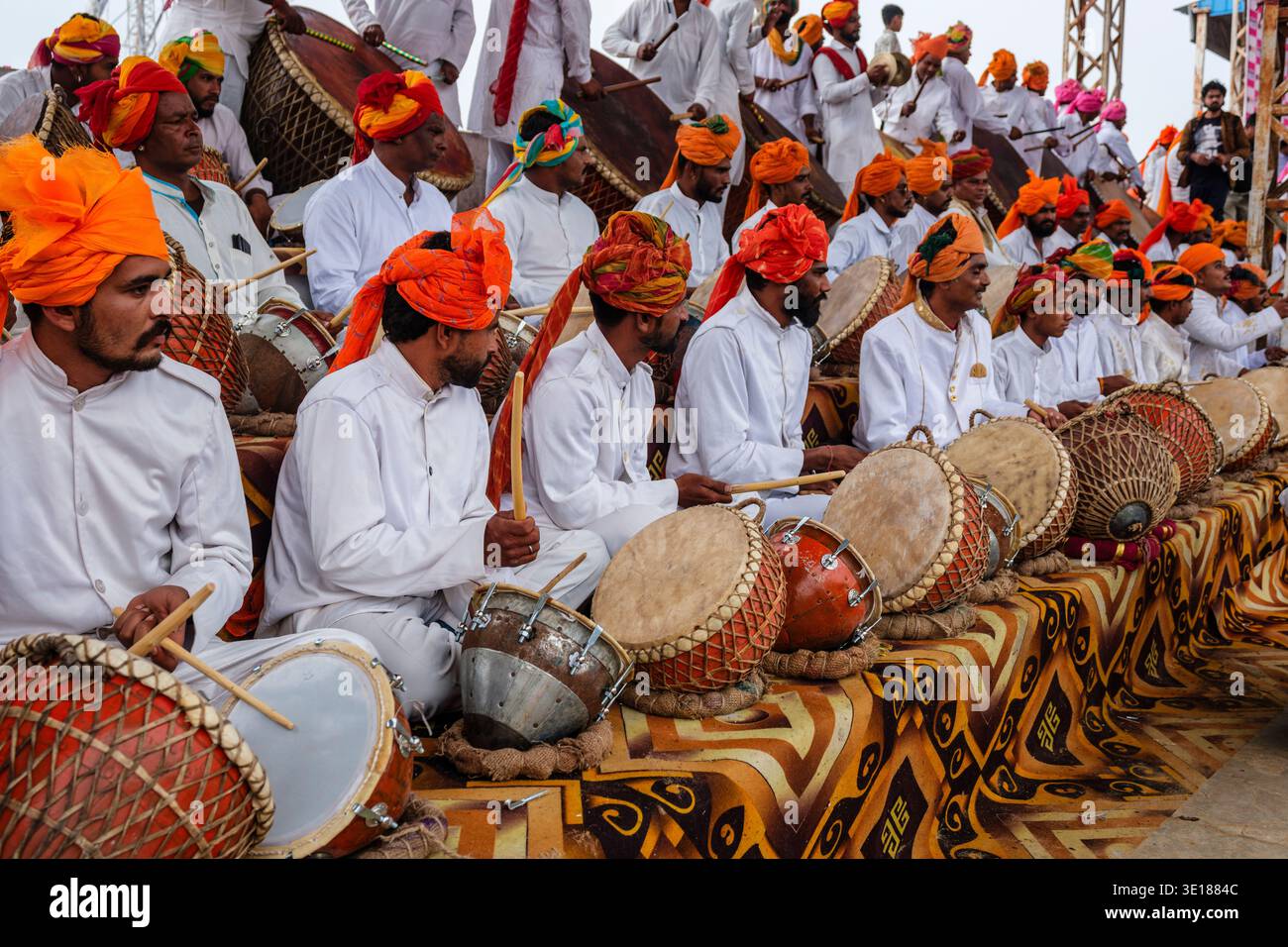 Grandi gruppi di uomini in uniforme bianca e turbanti arancioni suonano tamburi tradizionali mentre sono seduti su un tappeto a motivi geometrici durante Pushkar Mela. Foto Stock