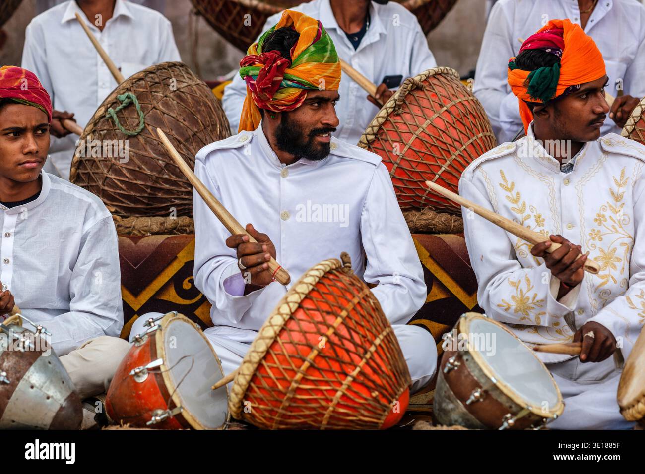 Un gruppo di musicisti al Pushkar Mela in camicie bianche e colorati turbanti si siedono insieme tenendo in mano bastoncini di legno e tamburi tradizionali. Foto Stock