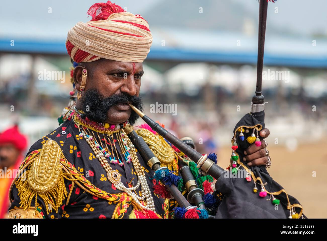 Un uomo con grandi baffi a Pushkar Mela indossa un'uniforme decorata e suona le cornamuse con una zona fieristica sfocata in lontananza. Foto Stock