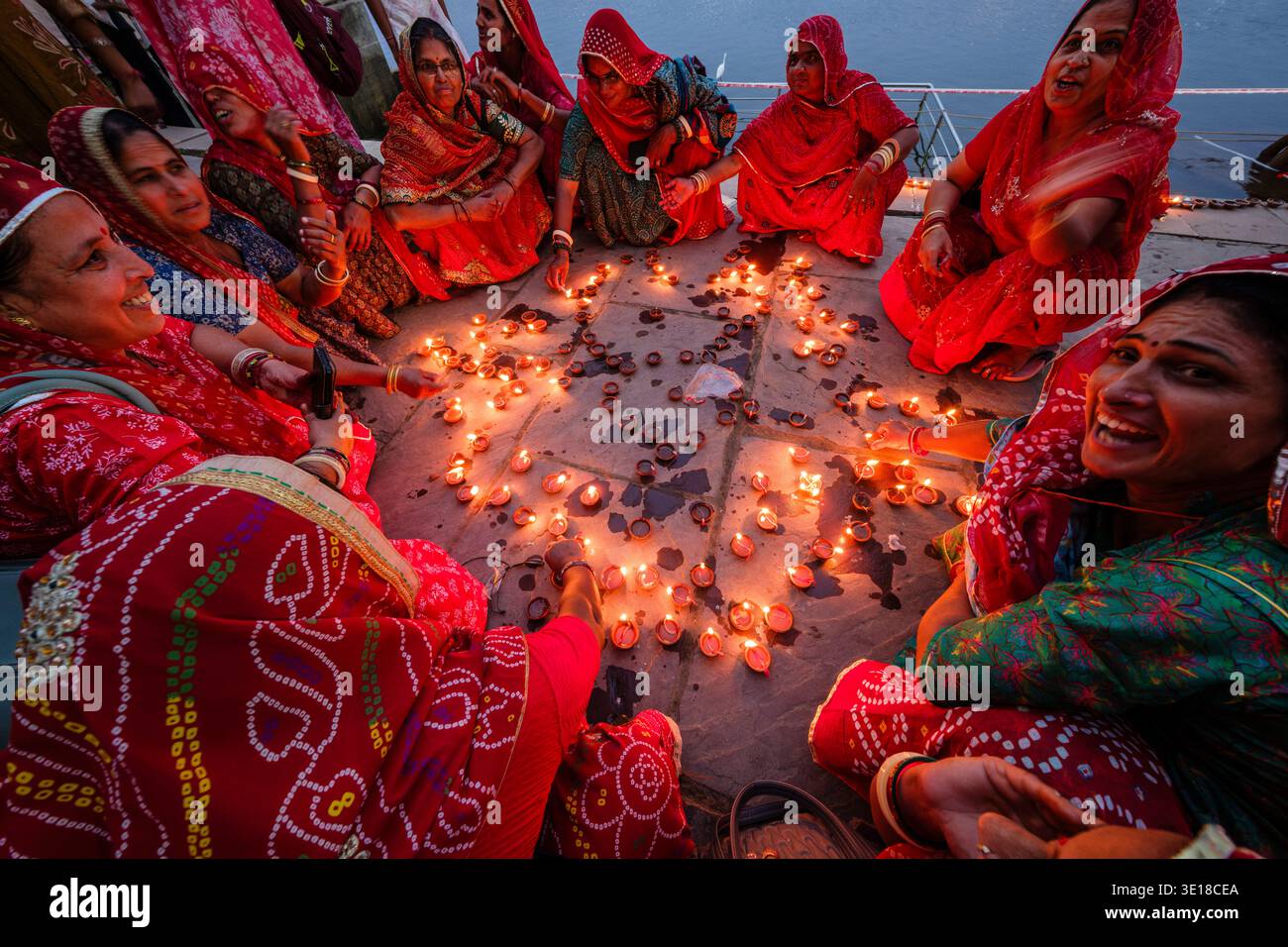 Un gruppo di donne in abito tradizionale siede accanto all'acqua al lago Pushkar, sistemando e accendendo numerose piccole lampade a olio sul terreno di pietra al crepuscolo. Foto Stock