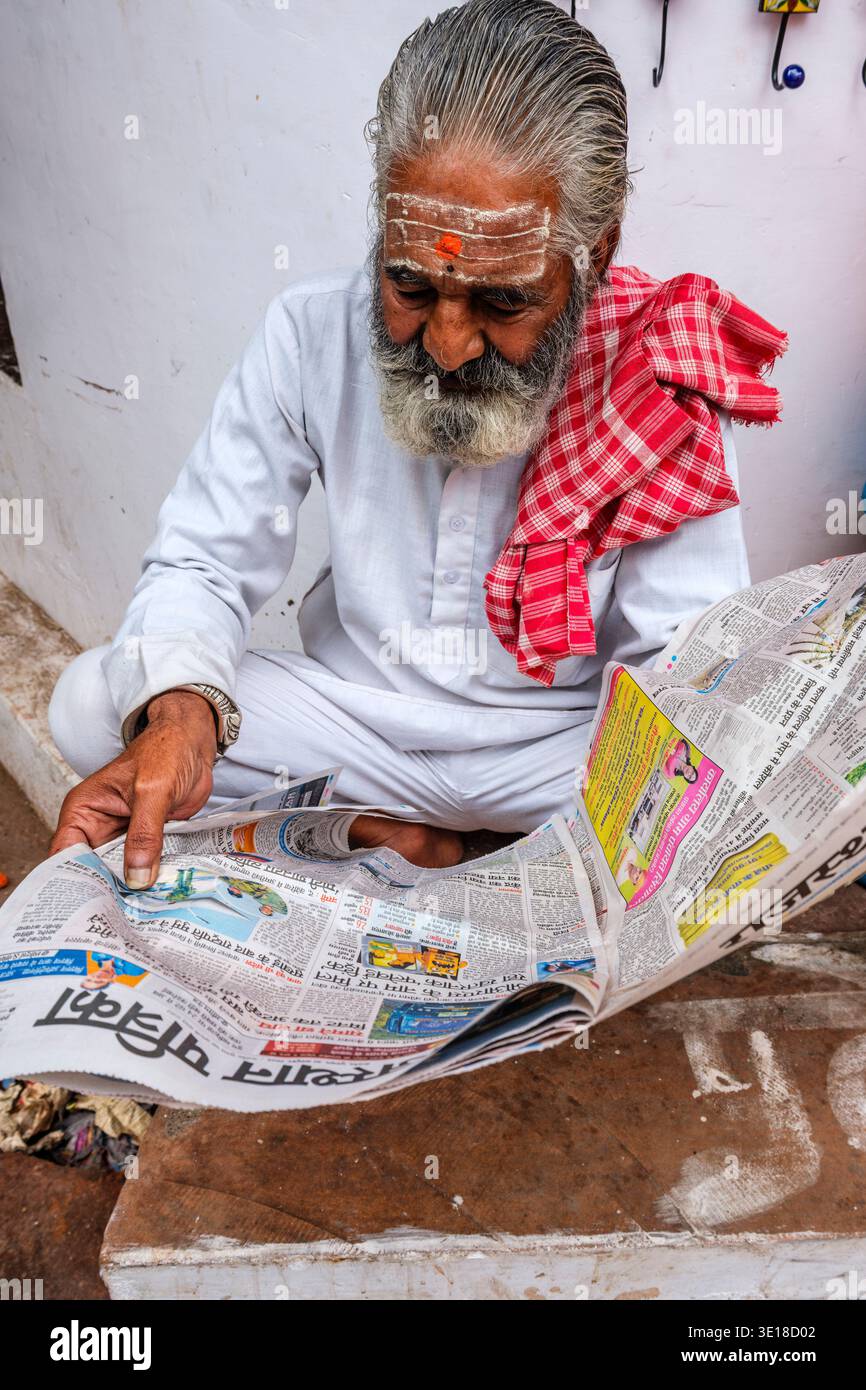Un uomo anziano con una barba grigia e un segno tradizionale sulla fronte si siede per terra a Pushkar mentre si concentra sulla lettura di un giornale. Foto Stock