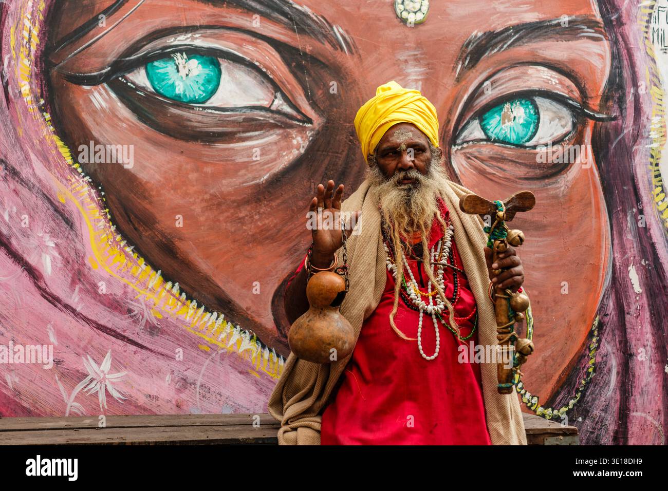 A Pushkar un Sadhu, un uomo sacro, in un turbante giallo e una veste rossa, regge un bastone di legno e una zucca, in posa davanti a un grande murale di occhi di una donna. Foto Stock