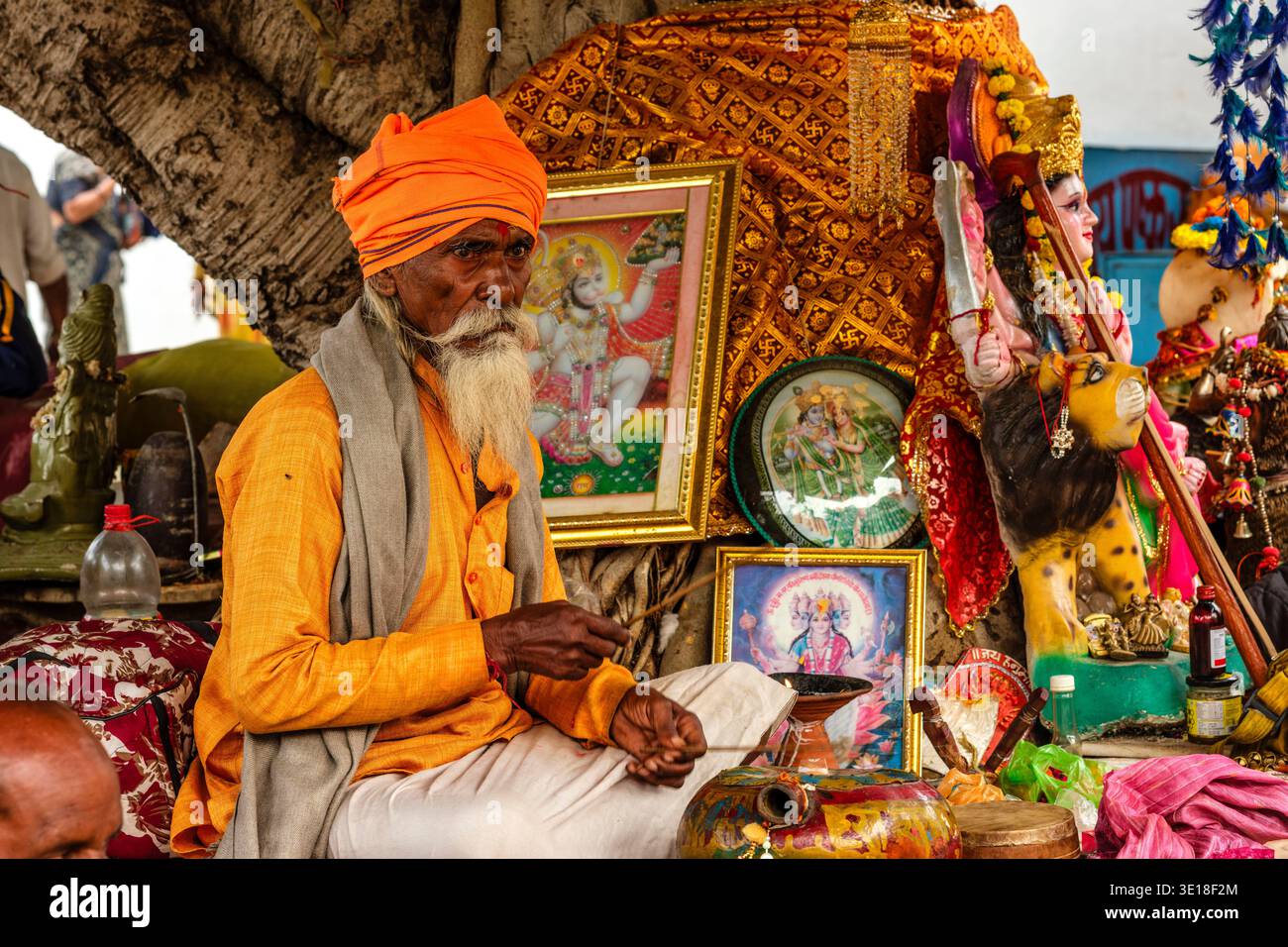 Un Sadhu o un santo uomo che indossa un turbante arancione si trova al Sunset Ghat presso il lago Pushkar, prima di una collezione di immagini incorniciate e piccole statue. Foto Stock