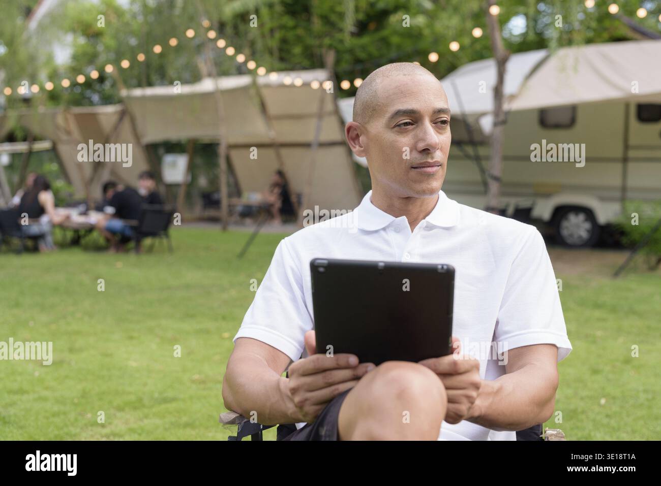 Elegante uomo calvo con una polo bianca che si rilassa in campeggio accanto a un camper utilizzando un tablet. Concetto di stile di vita per viaggi all'aperto e campeggio Foto Stock
