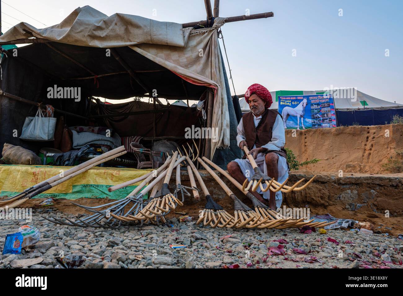Un venditore che indossa un turbante rosso assembla forchette di legno fuori dal suo rifugio temporaneo a Pushkar Mela, mentre un poster di cavallo è appeso in lontananza. Pu Foto Stock