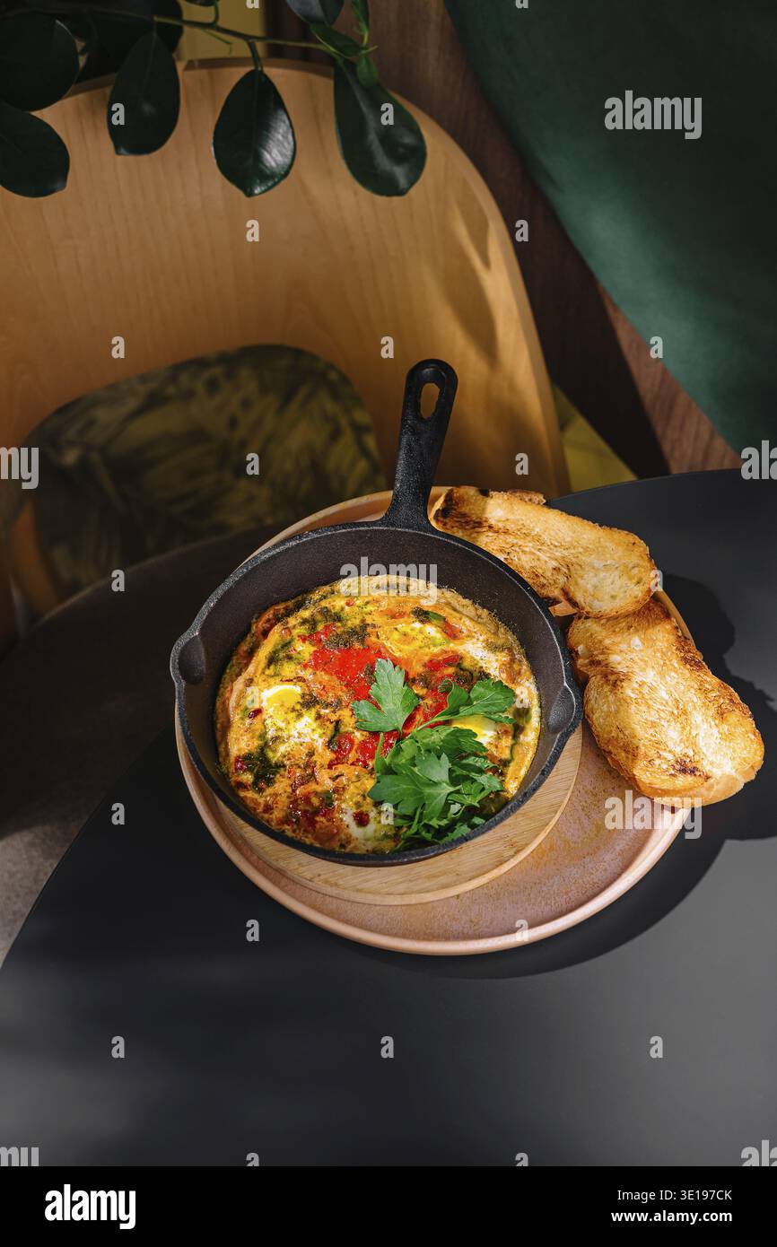Uova appena cucinate con verdure ed erbe aromatiche servite su vassoio di legno con pane tostato su un tavolo nero nel caffè Foto Stock