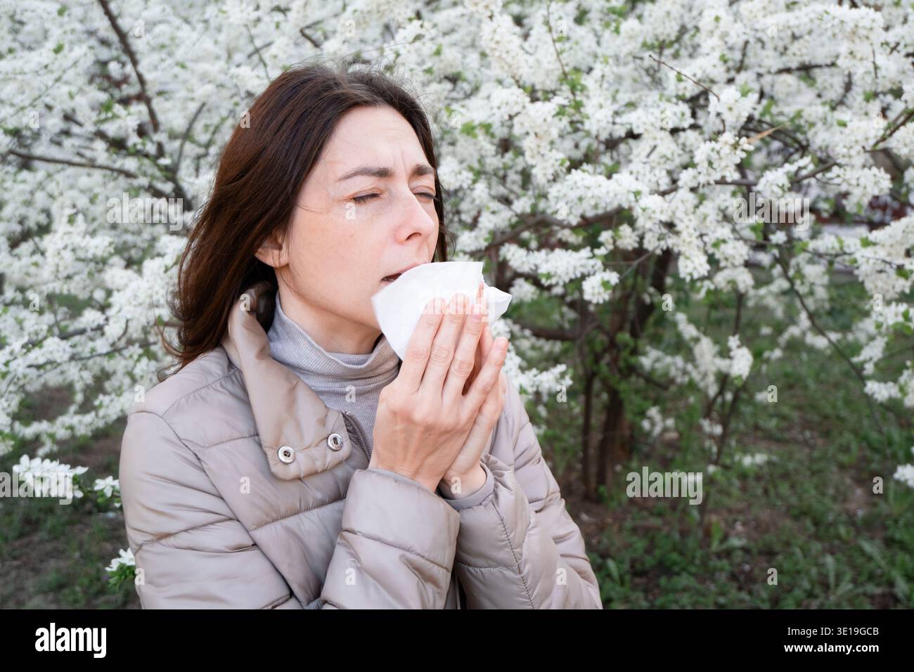 Donna che soffia il naso nel tessuto vicino ai fiori bianchi della primavera. Raffreddore stagionale o allergia, sintomi di febbre da fieno. Foto Stock