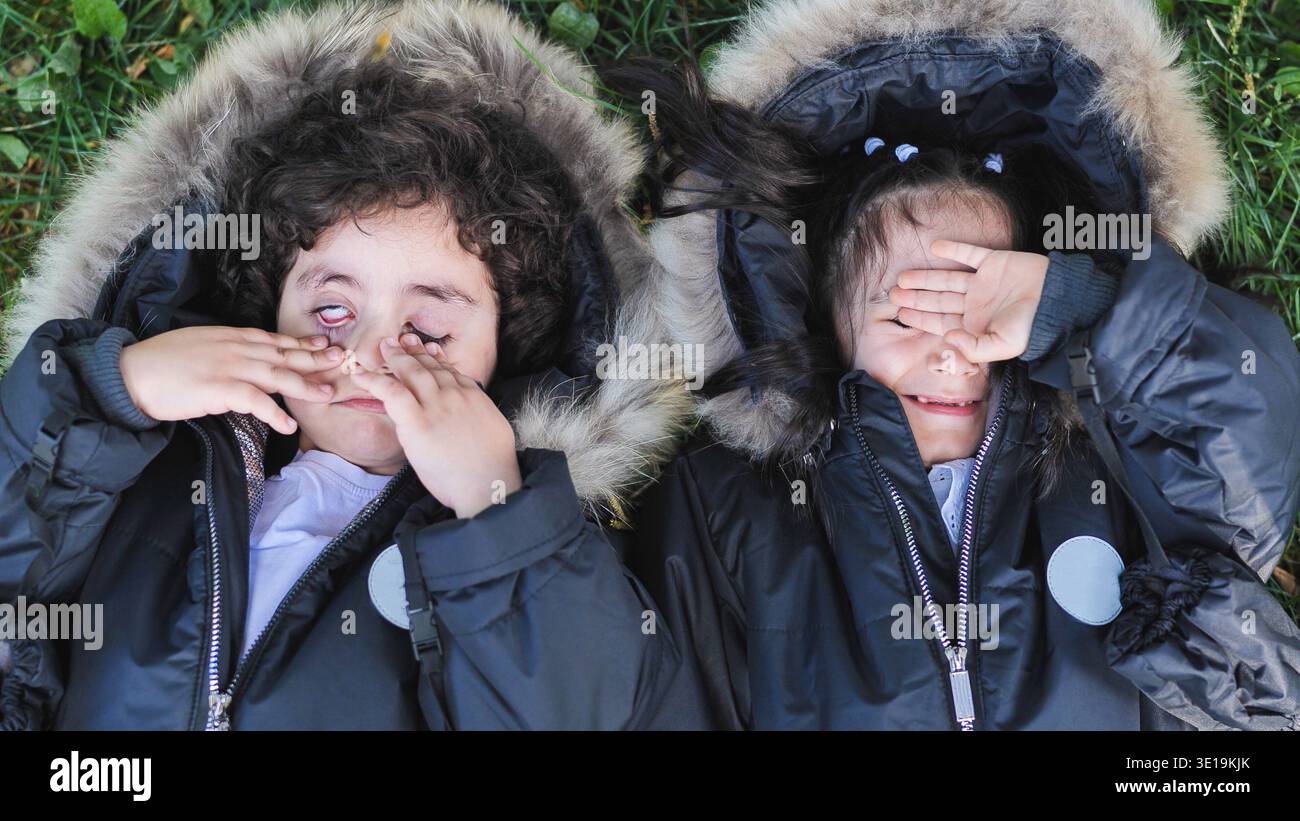 Bambini che fanno facce sciocche giocano all'aperto sull'erba con le calde giacche invernali Foto Stock