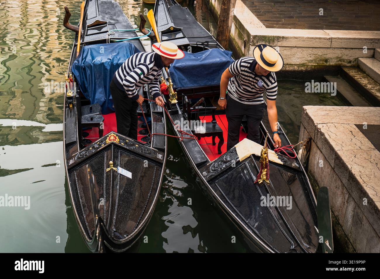 Gondolieri veneziani in camicie tradizionali a righe che preparano le loro gondole nere su un canale. Venezia, Italia. Foto Stock