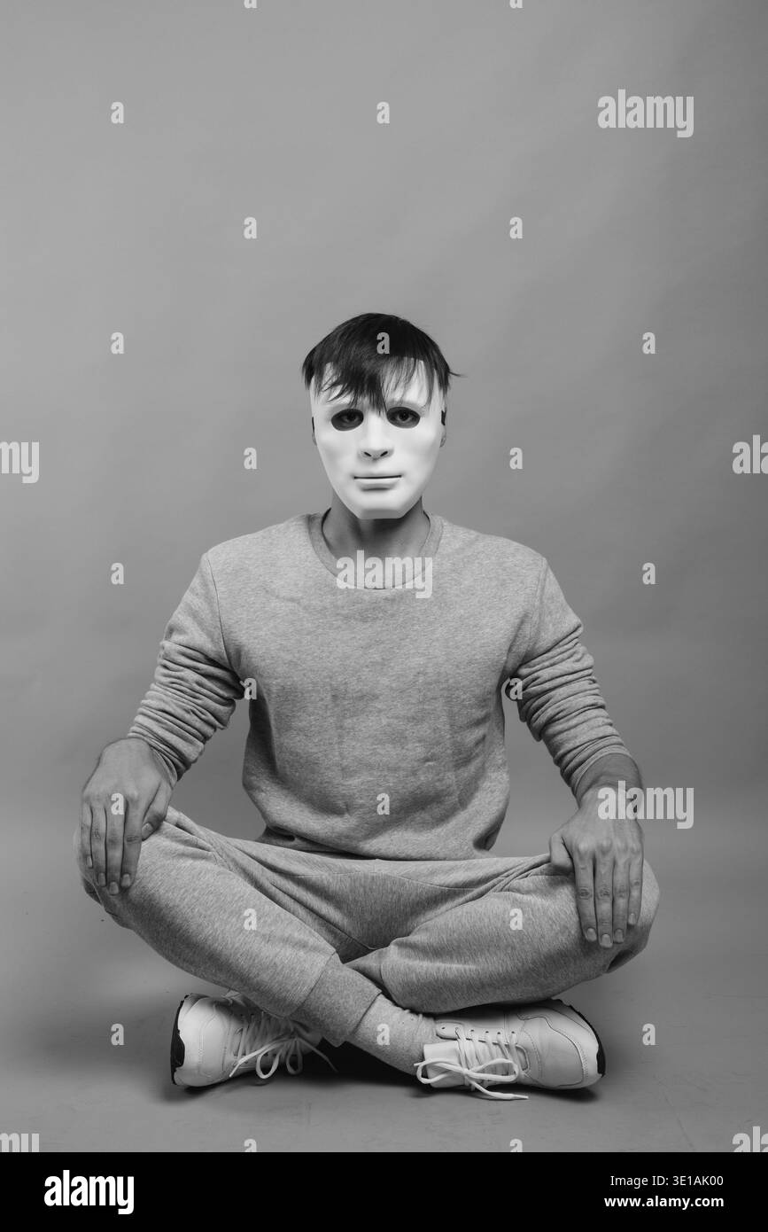 Studio shot del giovane uomo che indossa maschera bianca seduti con le gambe incrociate contro uno sfondo grigio Foto Stock