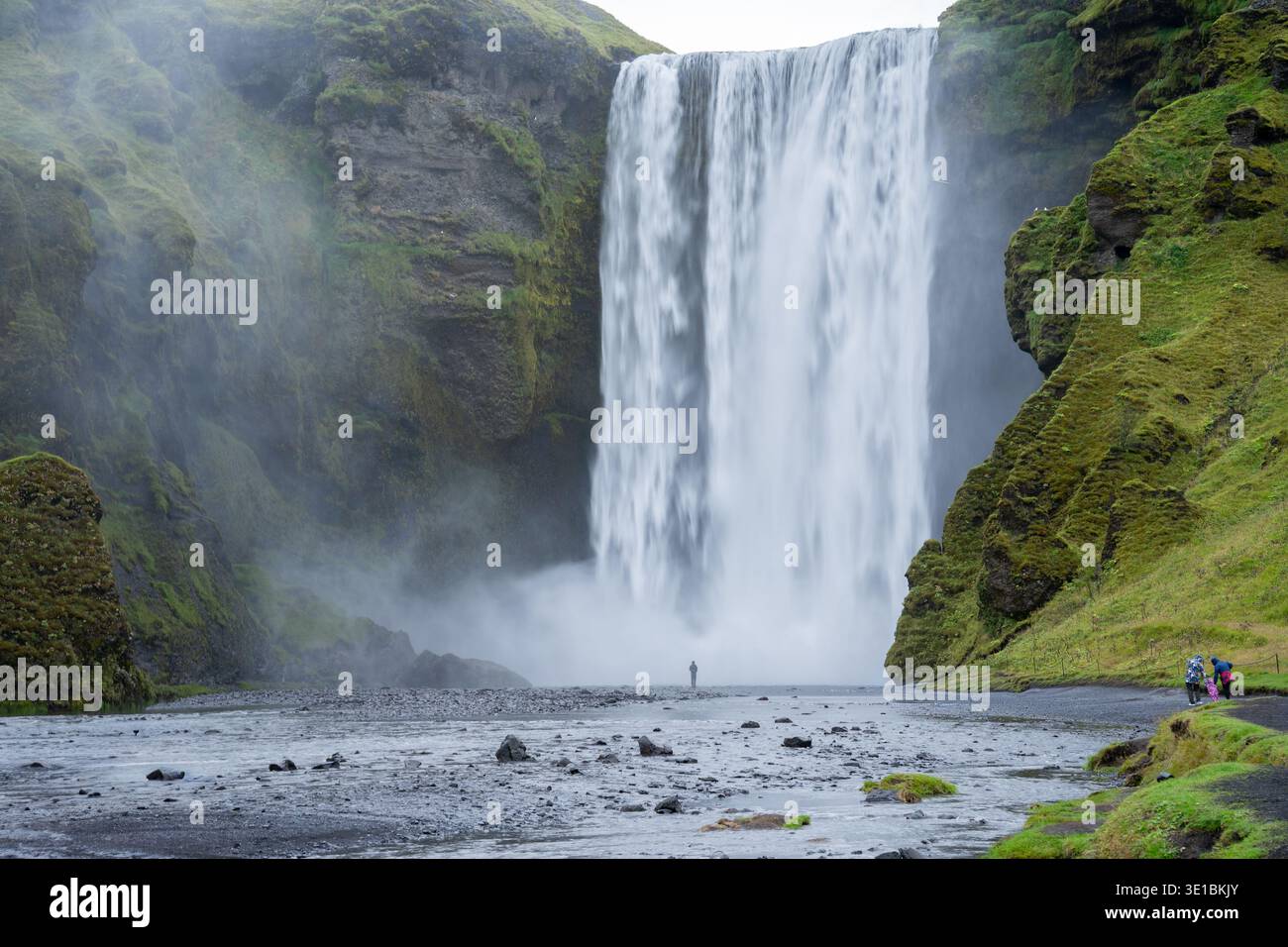 Enorme cascata circondata da scogliera rocciosa rocciosa e persone che camminano verso di essa, l'Islanda Foto Stock