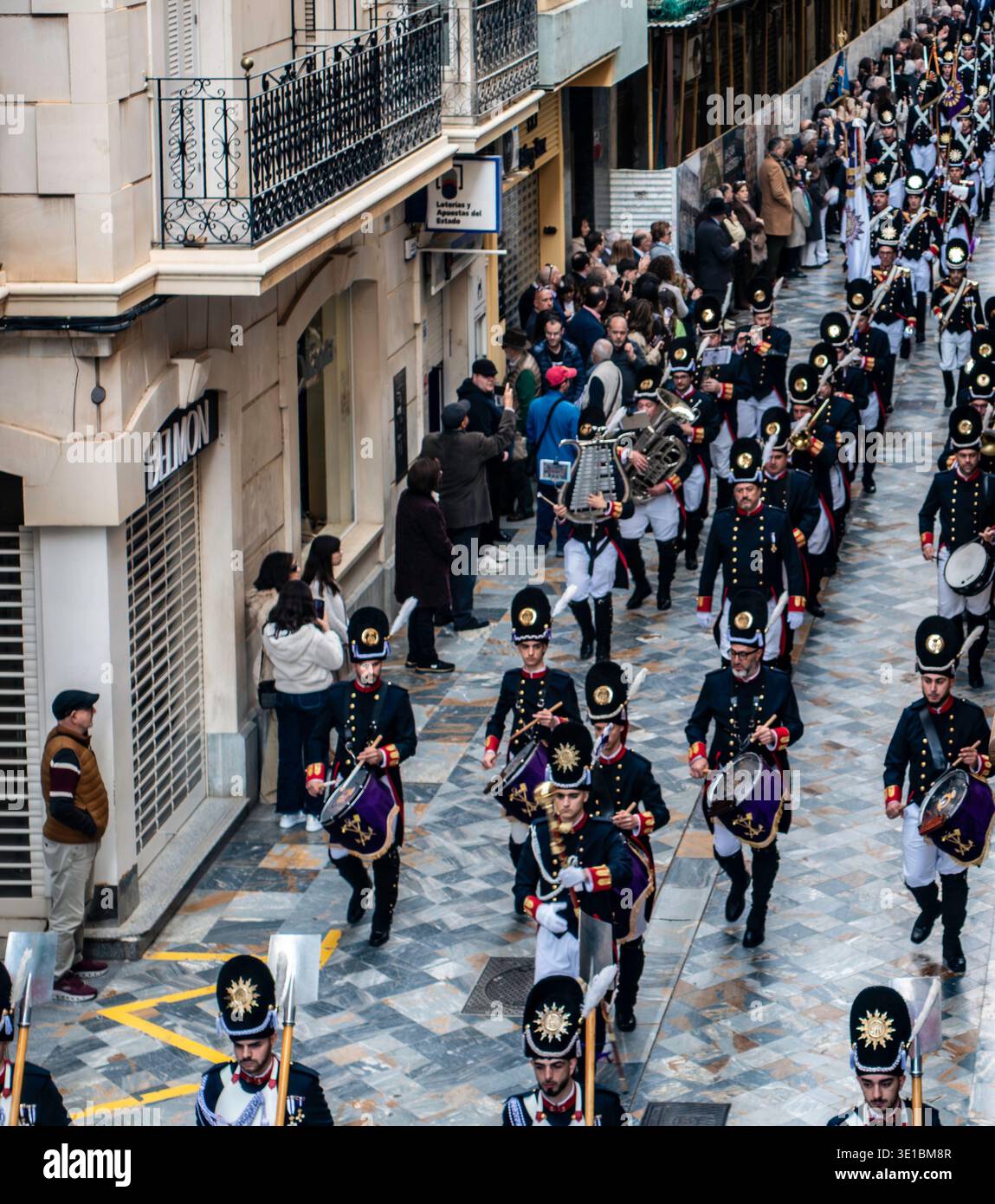 Una banda di marcia in uniforme nera con i cappelli shako sfilano lungo Calle Mayor a Cartagena, Spagna. Balconi ornati e facciate storiche fiancheggiano la strada Foto Stock