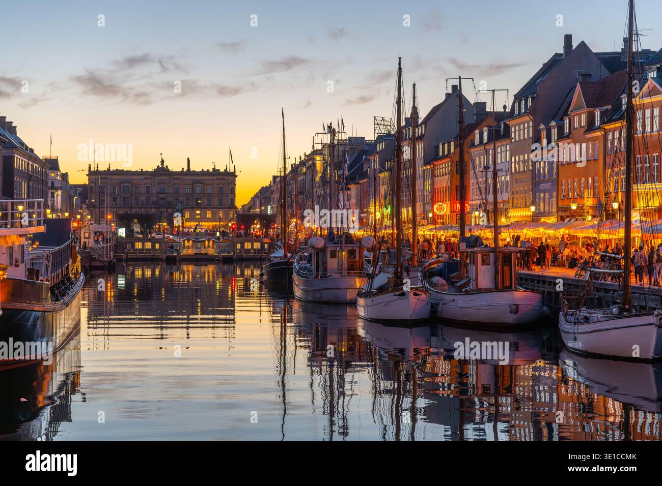 Mentre la sera scende su Nyhavn a Copenaghen, i colorati edifici illuminano il lungomare. Le barche si spostano dolcemente nel canale, mentre la gente si gode l'atmosfera vivace di accoglienti caffetterie. Foto Stock