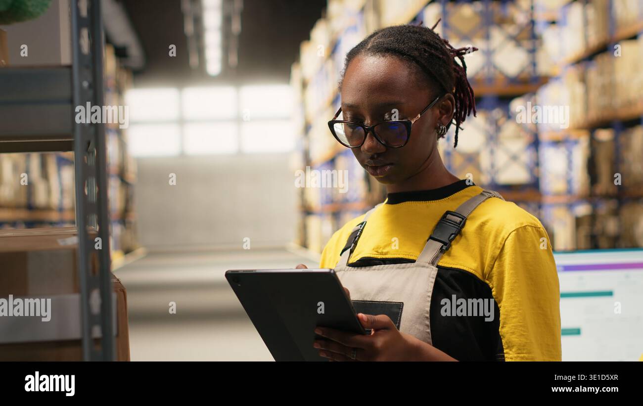 Una donna nera impiegata nell'elaborazione degli ordini con un tablet, il centro di distribuzione garantisce un funzionamento efficiente del magazzino. Gestione dei dati relativi ai pacchi per la consegna tramite e-commerce. Telecamera B.. Foto Stock