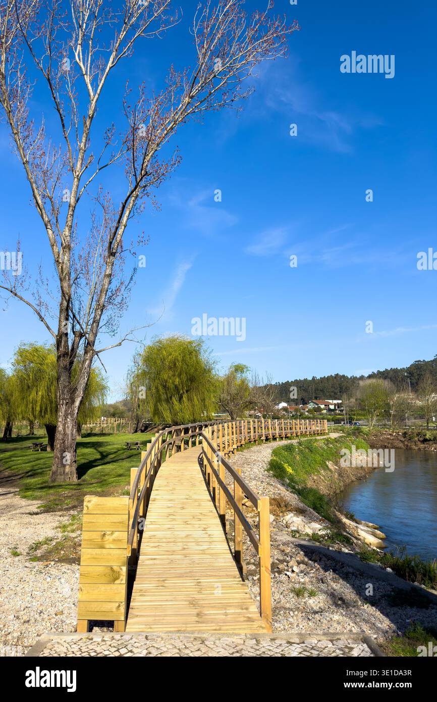 Una passerella in legno conduce l'occhio attraverso il paesaggio panoramico del Parque Municipal do Antua. La composizione presenta alberi primaverili in erba, un fiume calmo, Foto Stock