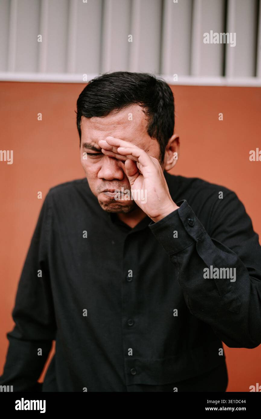 Un uomo con una camicia nera si strofina gli occhi, esprimendo disagio, stanchezza o mal di testa, probabilmente a causa dello sforzo oculare degli schermi o della mancanza di sonno. Foto Stock