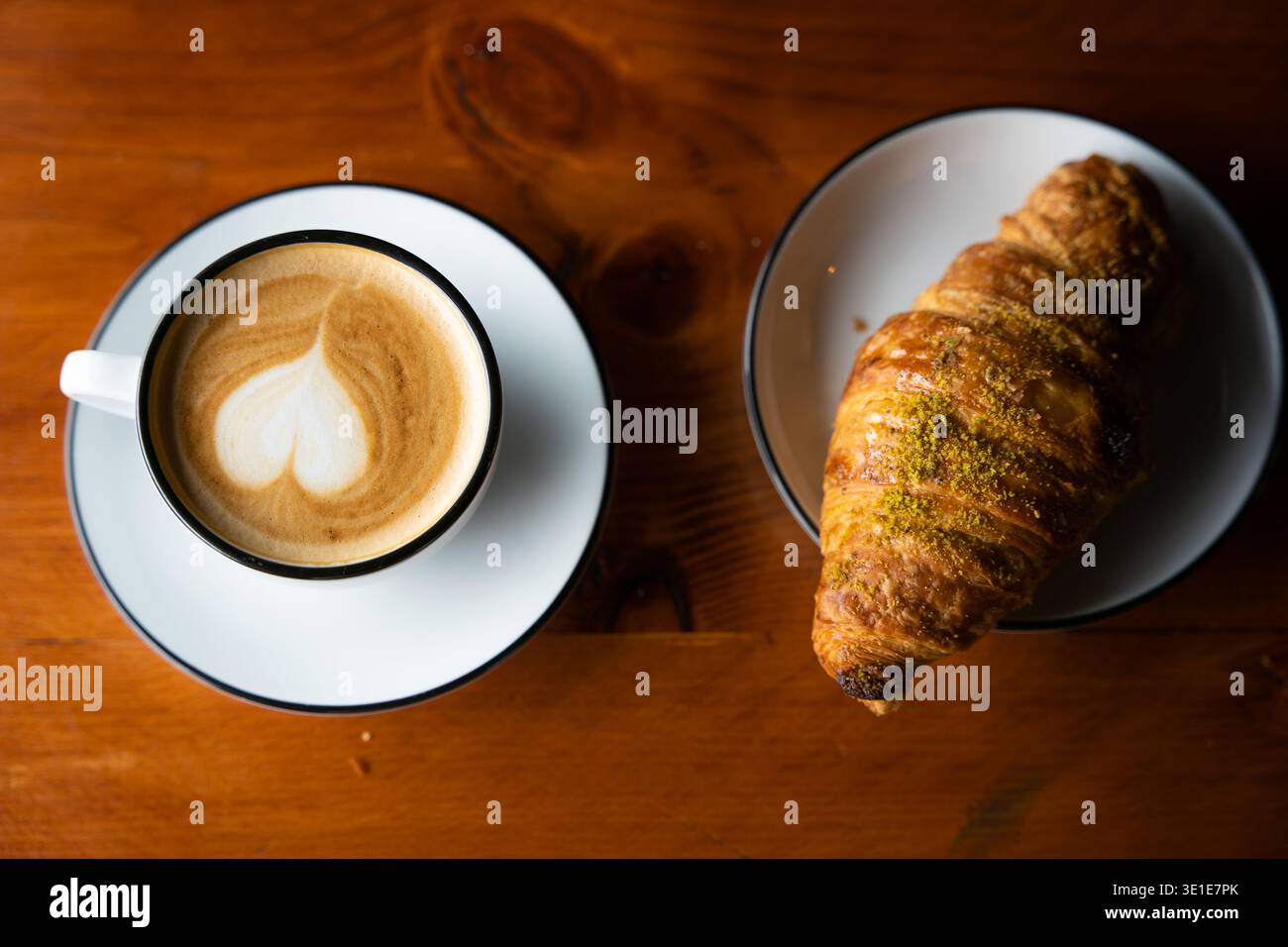 Una tazza di caffe' latte con intricate opere d'arte in schiuma si trova accanto a un croissant dorato su un tavolo da caffe' in legno in Tunisia. Foto Stock