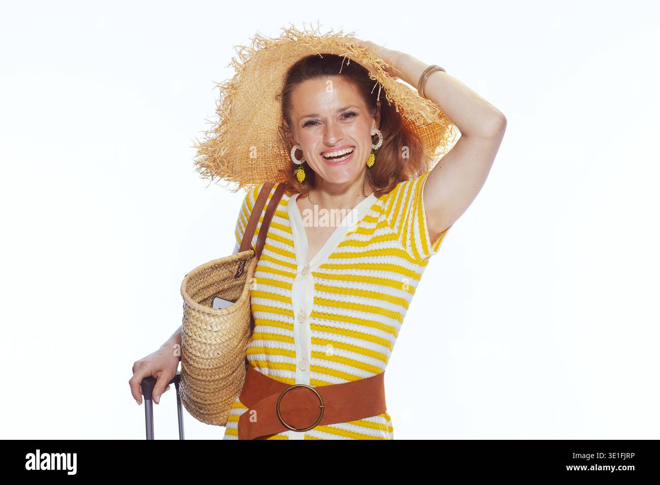 Una bella donna con un abito estivo a righe e un cappello a tesa larga sorride tenendo in mano la borsa di paglia e il manico della valigia, che incarna lo stile delle vacanze. Foto Stock