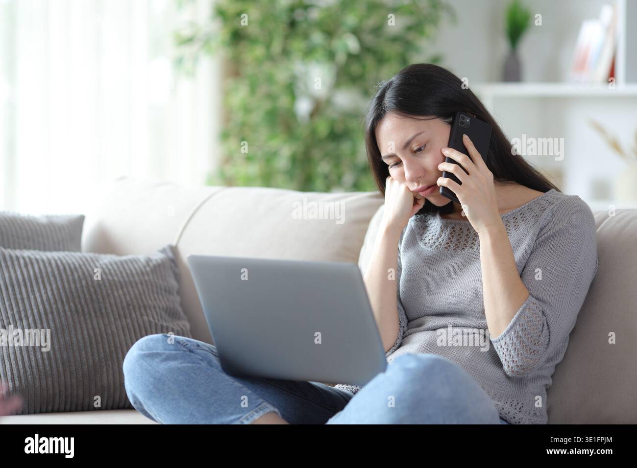 Annoiata donna asiatica che chiama al telefono controllando il laptop seduto su un divano a casa Foto Stock