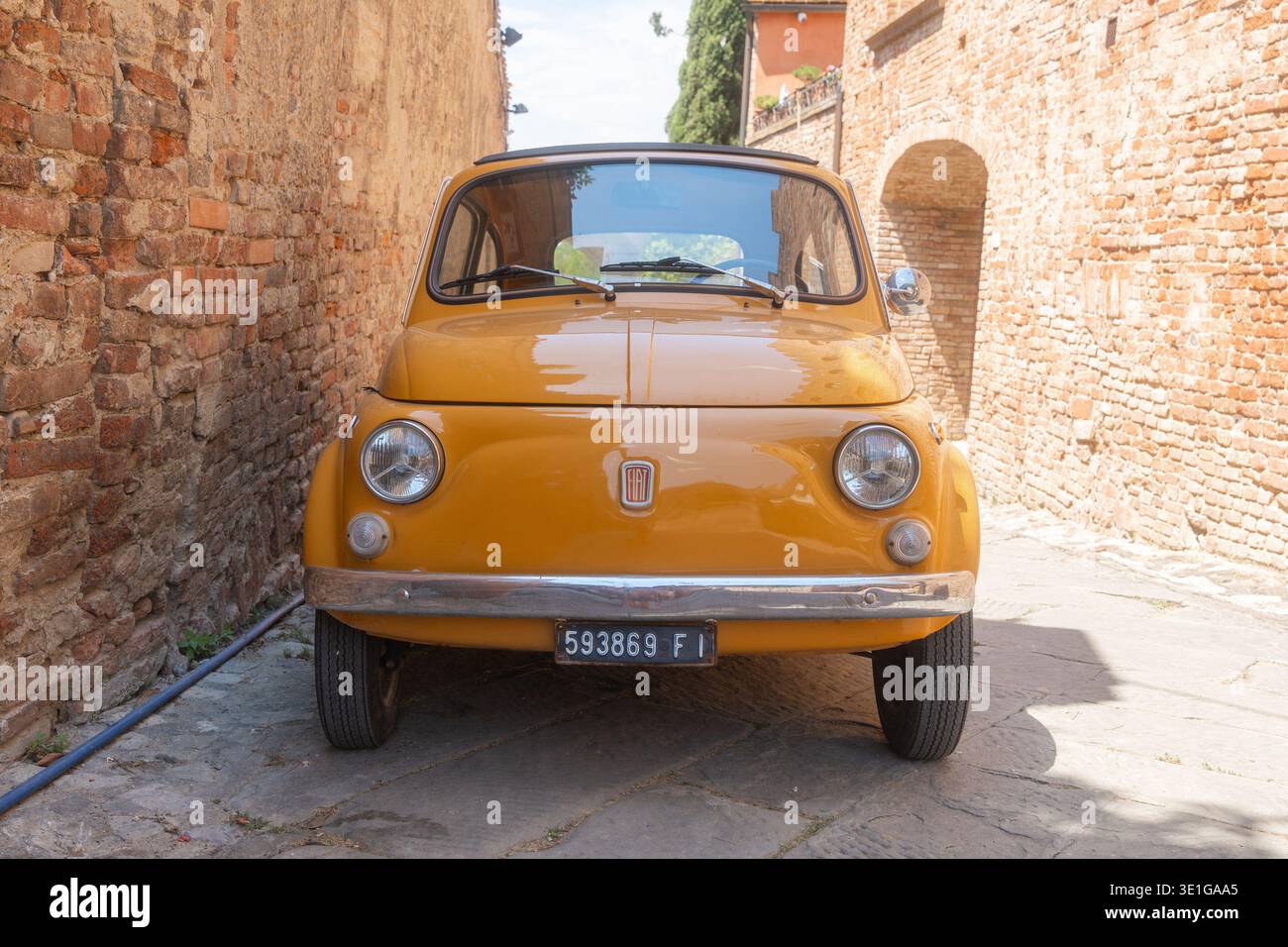 Baccoleno, Italia, 05.07.2026, auto d'epoca gialla parcheggiata in una stretta strada di pietra di un centro storico europeo. Luce calda, pareti in mattoni testurizzati e. Foto Stock
