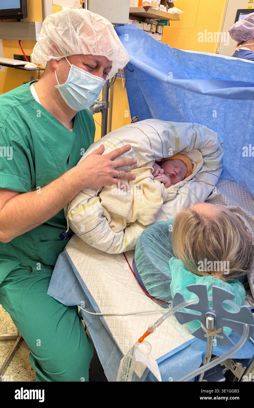 Padre che tiene in braccio il neonato in sala operatoria mentre la madre si trova sul tavolo chirurgico durante il taglio cesareo. Momento emotivo del primo contatto Foto Stock
