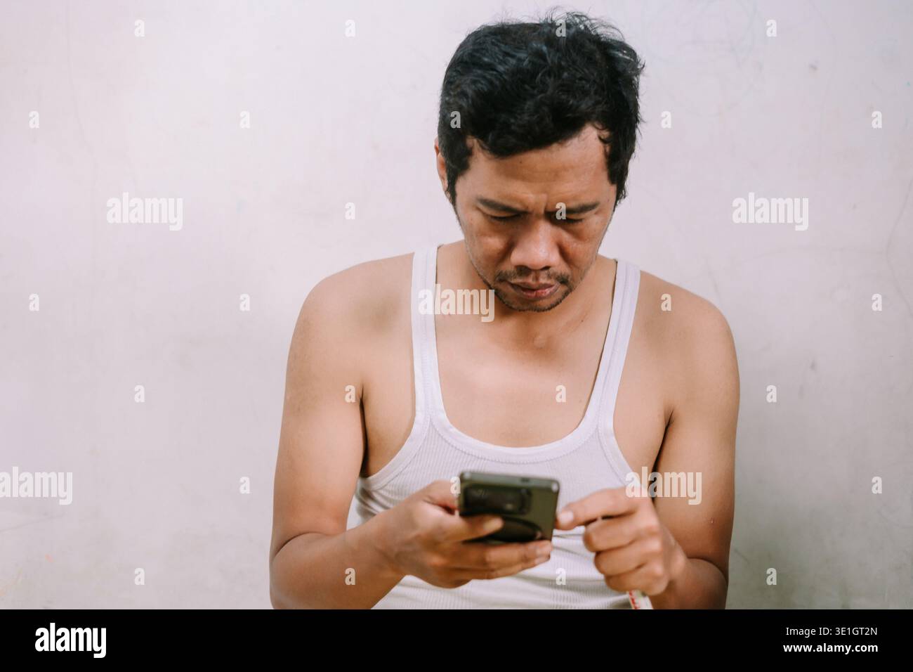 Un uomo con una canotta bianca è incentrato sul suo smartphone. La sua espressione seria e concentrata suggerisce che sta leggendo messaggi importanti o. Foto Stock