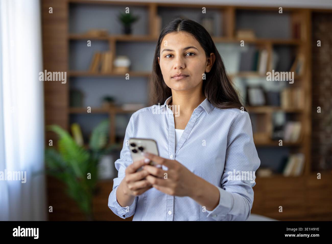 Giovane donna in un ufficio domestico, in possesso di uno smartphone, che rappresenta la comunicazione moderna, il lavoro online e la tecnologia digitale in uno spazio di lavoro remoto Foto Stock