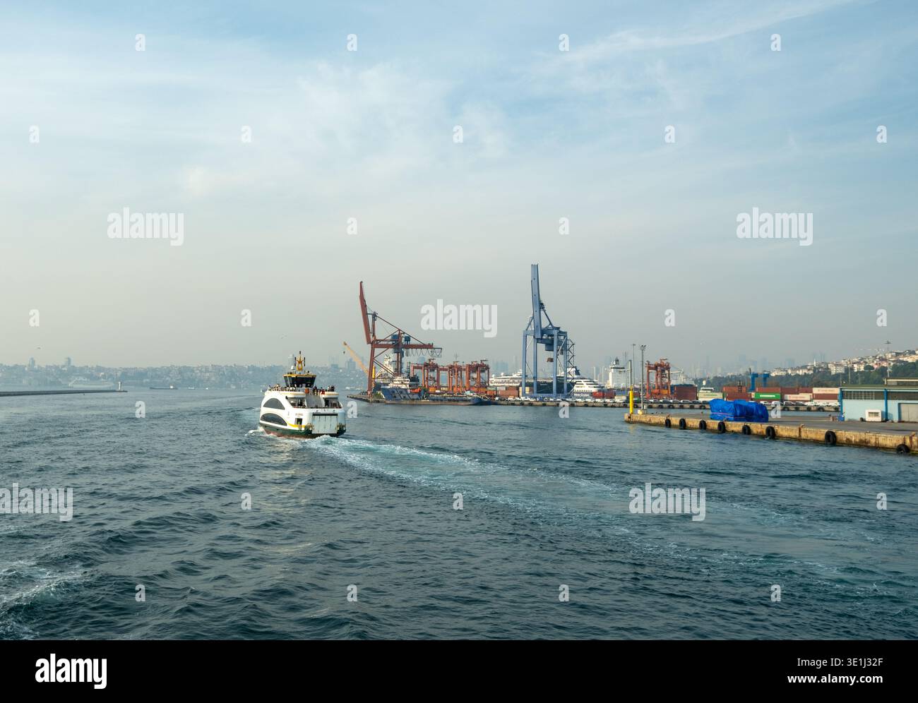 Il traghetto bianco passa davanti al porto industriale per container con colorate gru e container. L'acqua blu crea un risveglio dietro il ferro Foto Stock