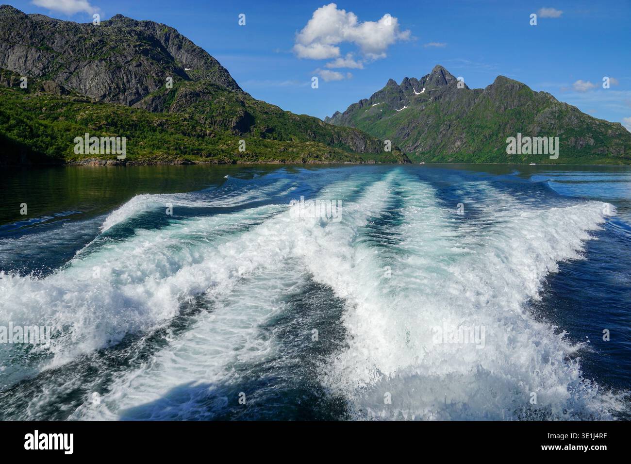 Il risveglio in barca si estende attraverso le calme acque del fiordo con ripide montagne e cielo blu sullo sfondo. Foto Stock