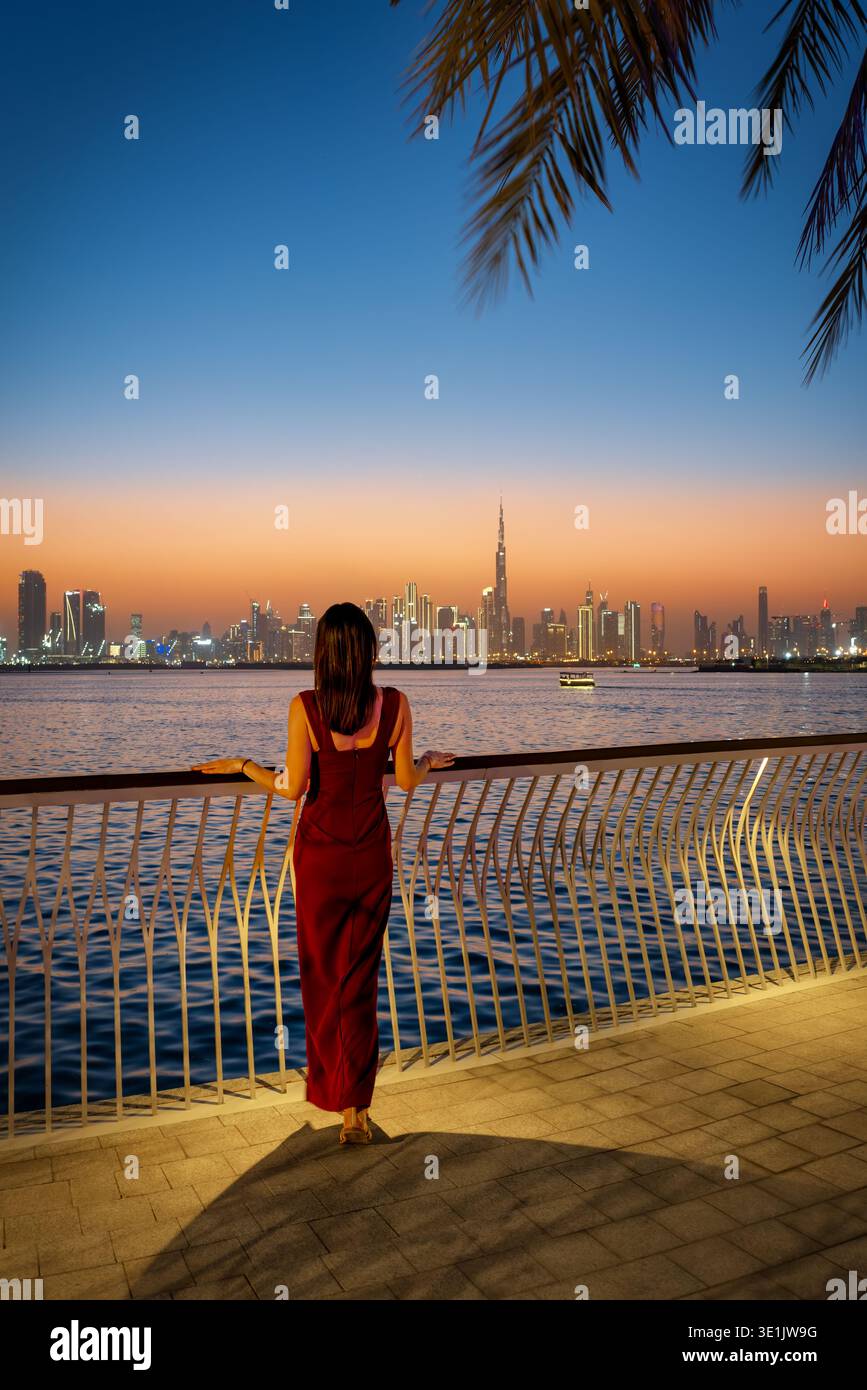 Una bella donna con un abito da sera rosso che guarda lo skyline illuminato di Dubai, Emirati Arabi Uniti, al crepuscolo dal porto di Creek Foto Stock