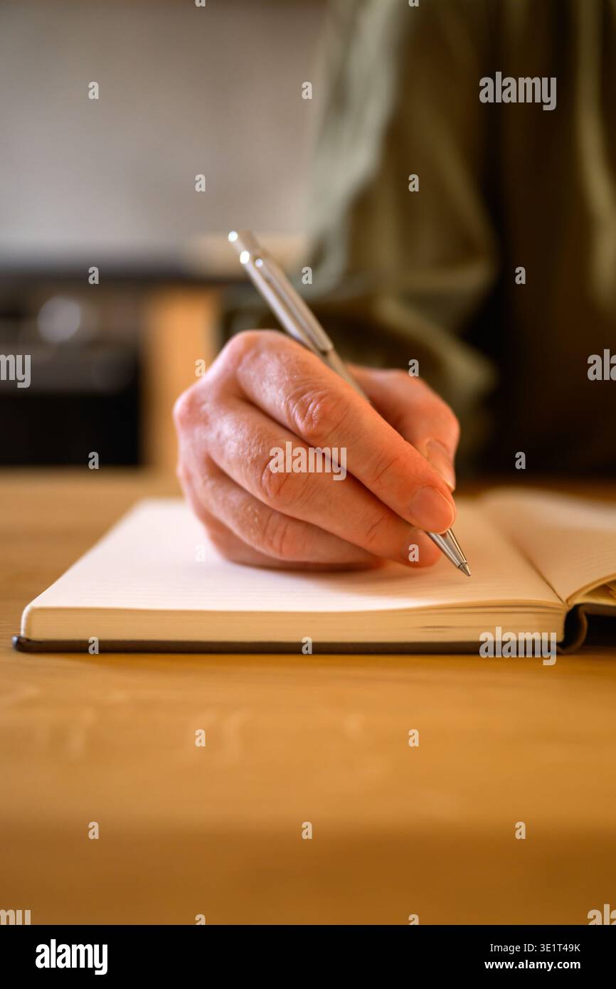 Primo piano di una donna che scrive appunti su un notebook alla scrivania in legno con penna, luce naturale calda, home Office o lavoro freelance. Messa a fuoco selettiva. Foto Stock