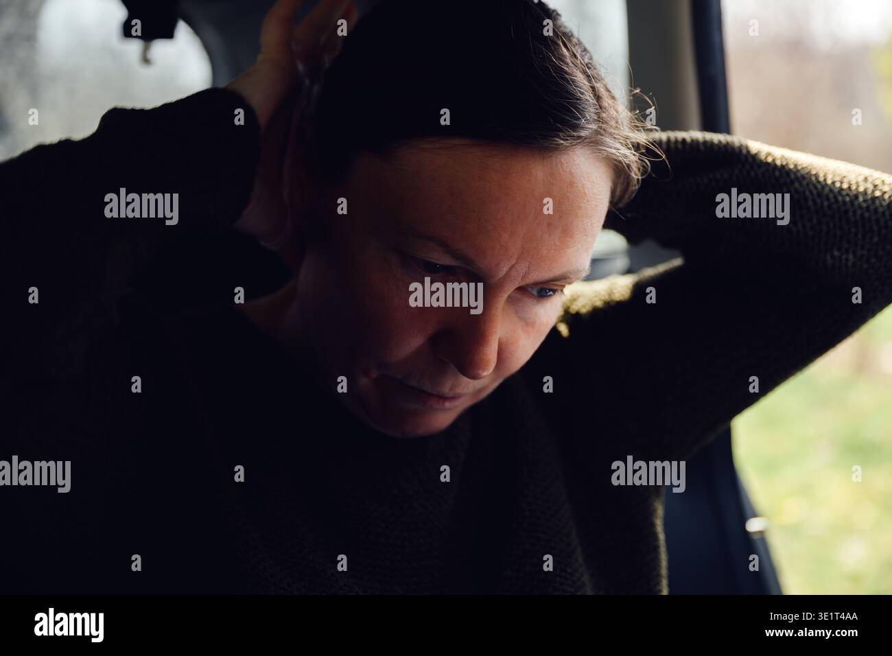 Donna che lega i capelli alla coda di cavallo seduto sul sedile posteriore dell'auto, illuminazione interna scura e soffusa, messa a fuoco selettiva Foto Stock