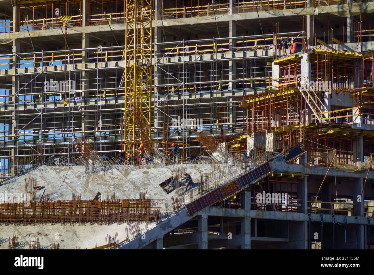 Operai che costruiscono un alto e moderno edificio, casseforme in cemento, ponteggi, gru a torre. Scena delle costruzioni industriali che mostra lavoro di squadra, equipaggiamento di sicurezza, sviluppo urbano. Foto Stock
