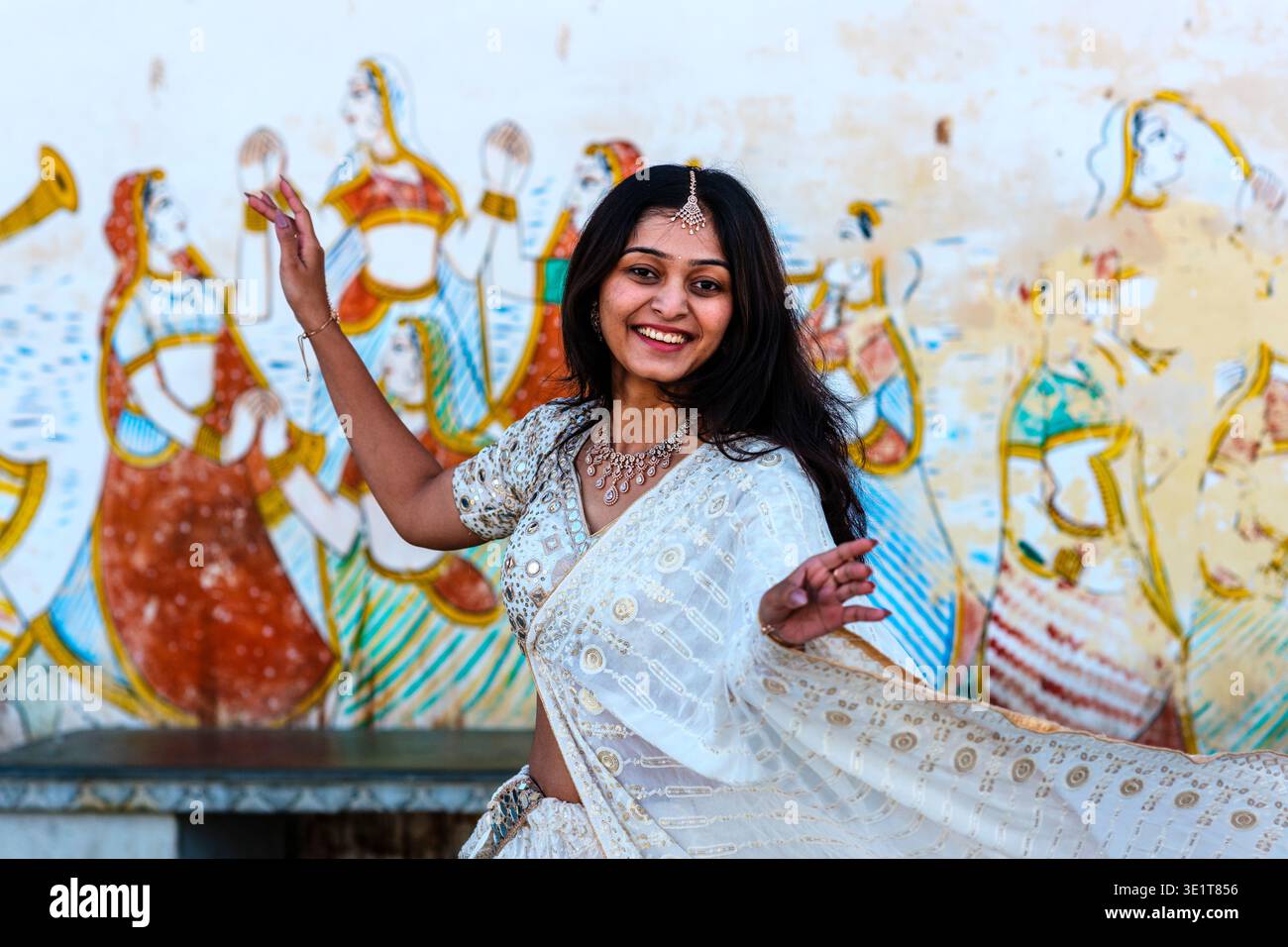 Una donna sorridente in una lehenga bianca posa a Udaipur davanti a un muro che presenta tradizionali dipinti Rajasthani di figure femminili e musicisti. Foto Stock