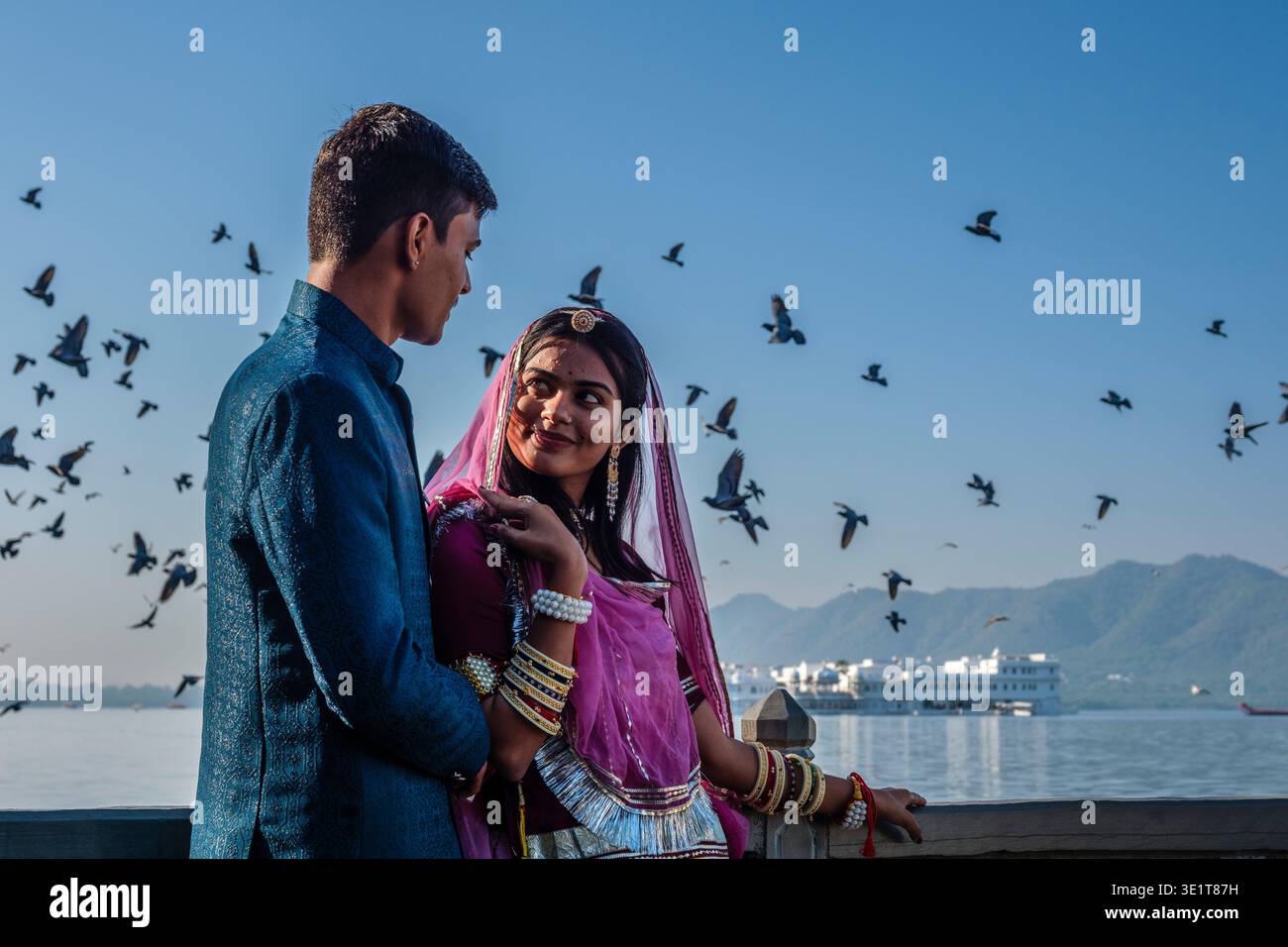 Una coppia in abbigliamento tradizionale si guarda l'un l'altro al lago Pichola a Udaipur, con piccioni e il Palazzo del Lago Taj visibili in lontananza. Foto Stock